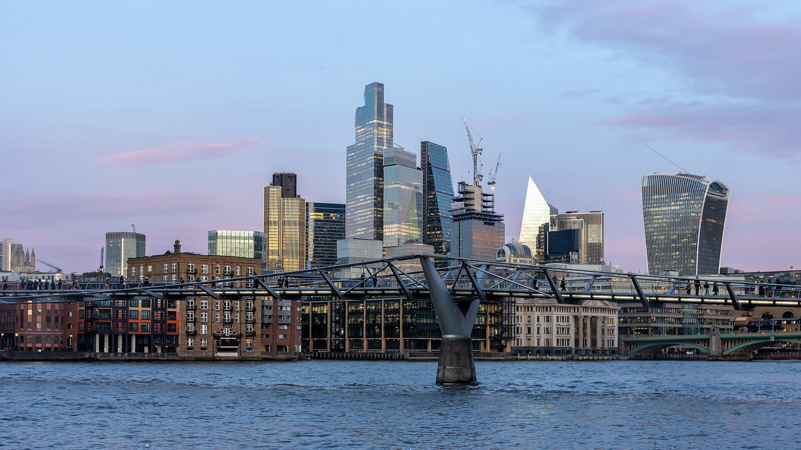 London Millennium Bridge in front of the City of London at dusk